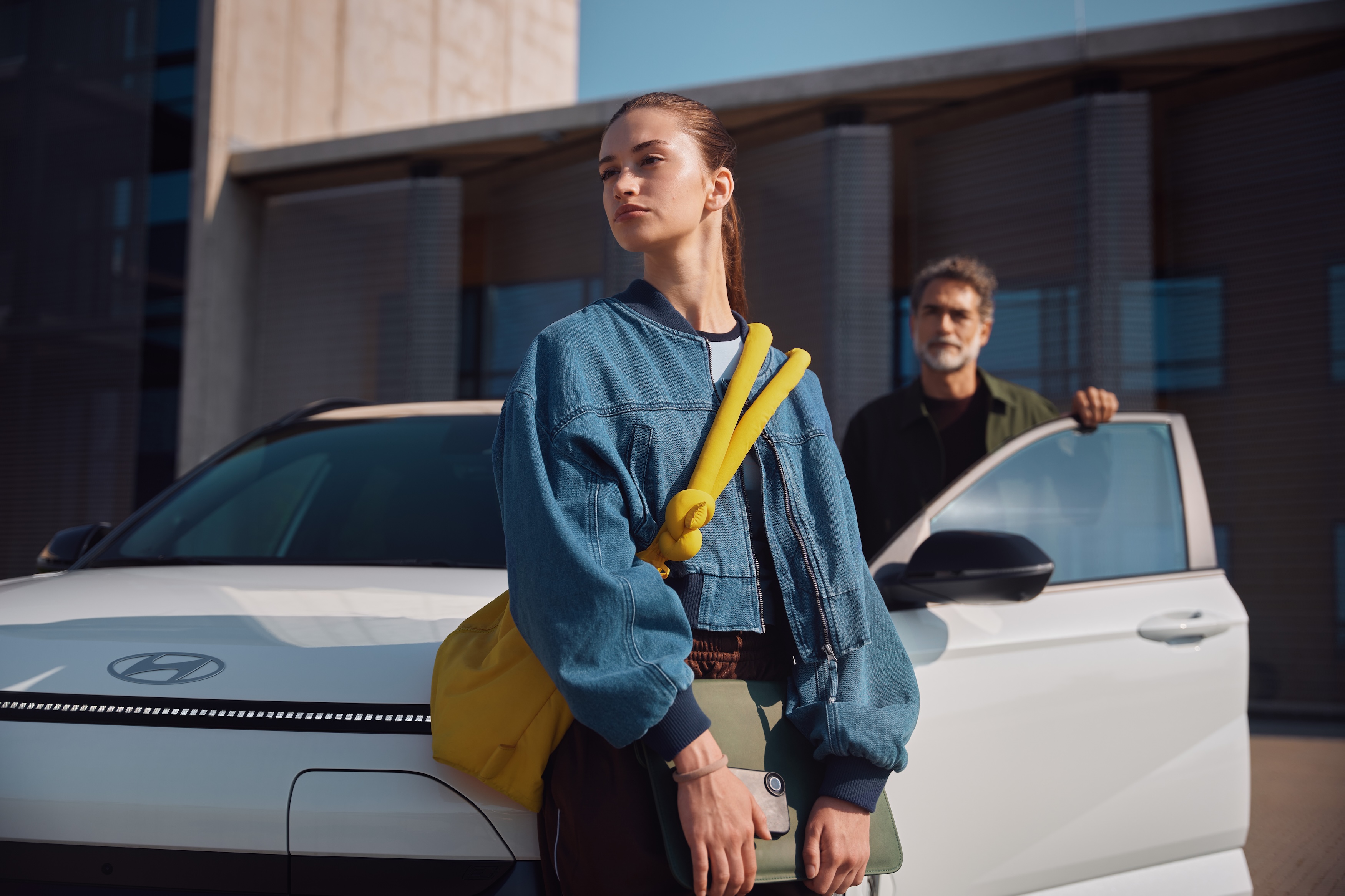 A young woman with a yellow bag leans against a white Hyundai Kona while a man is visible in the car's open door behind her. The scene is outdoors in front of a modern building.
