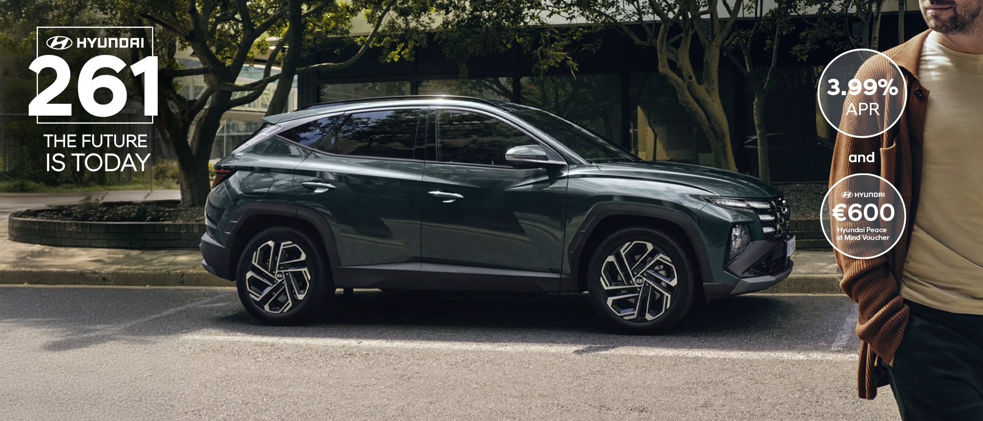 A man walking near a Hyundai TUCSON Hybrid parked by some trees.